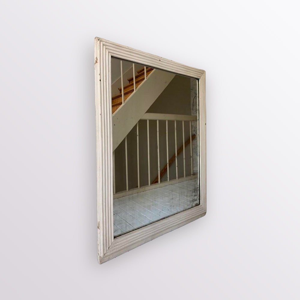 A square wall mirror with a distressed, weathered off-white wooden frame, mounted on a plain white wall. The mirror reflects a staircase with wooden railings and light-colored flooring.