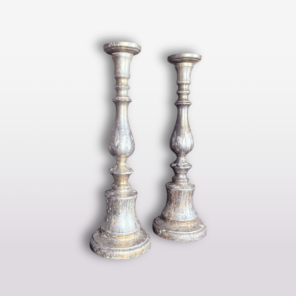 A pair of distressed silver-toned metal candlesticks with a weathered, vintage finish, standing upright against a light gray gradient background.