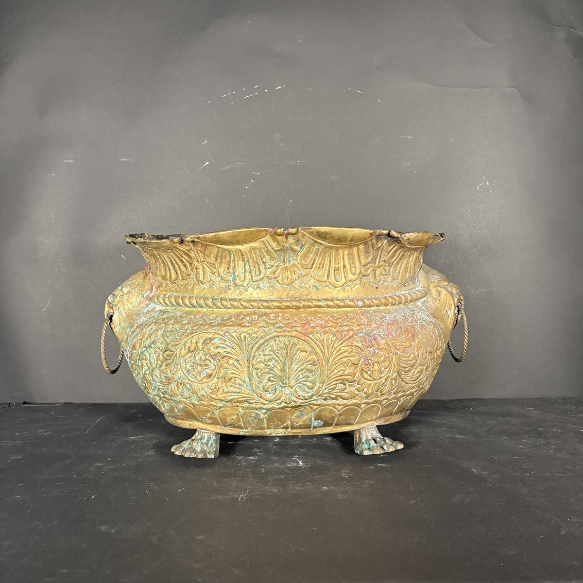 An antique bronze-colored metal planter or bowl with embossed floral patterns and side loop handles, displayed on a dark surface against a dark gray background.