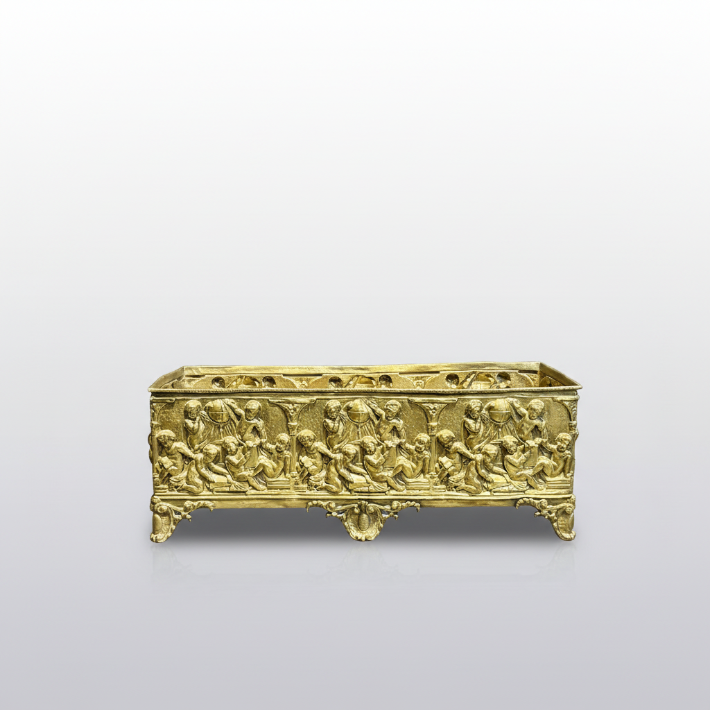 An ornate rectangular gold-toned metal box with intricate relief carvings depicting figures, standing on a dark gray surface against a dark gray backdrop.