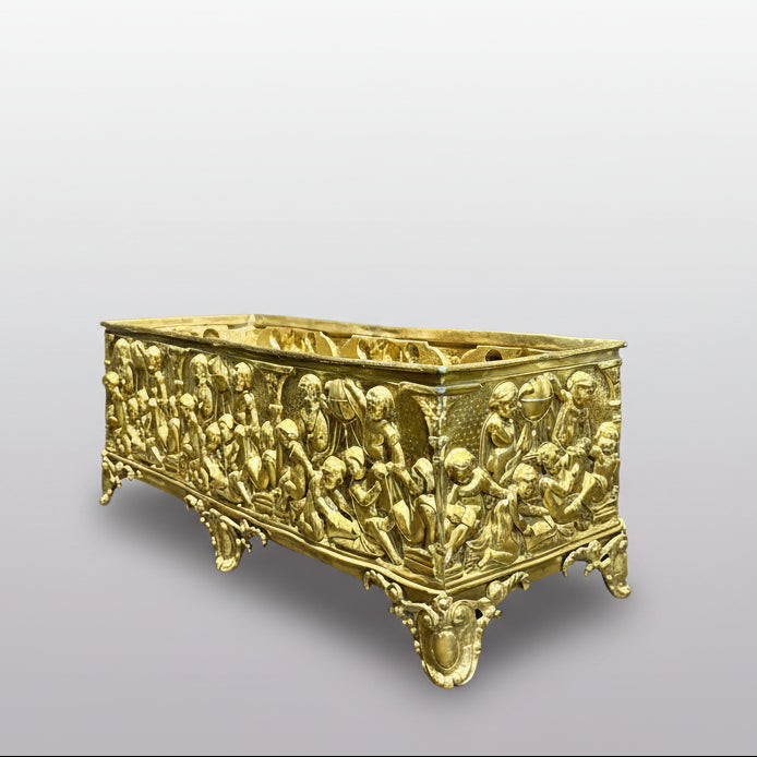 An ornate rectangular gold-toned metal planter with intricate relief carvings of classical figures, displayed on a gray surface against a dark gray backdrop.