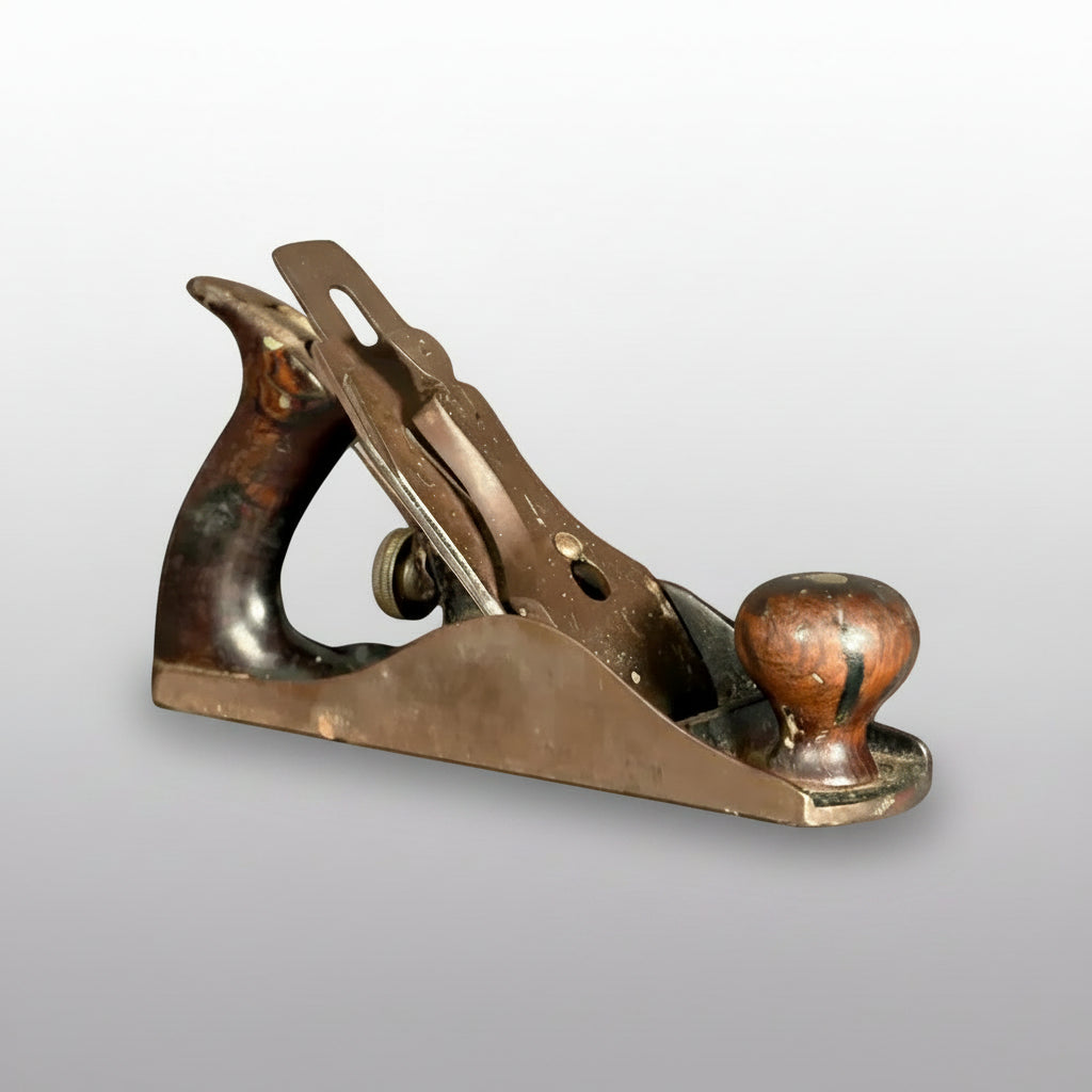 A vintage metal hand plane with a dark brown wooden handle and a copper-toned metal body, resting on a gray surface against a gray background.
