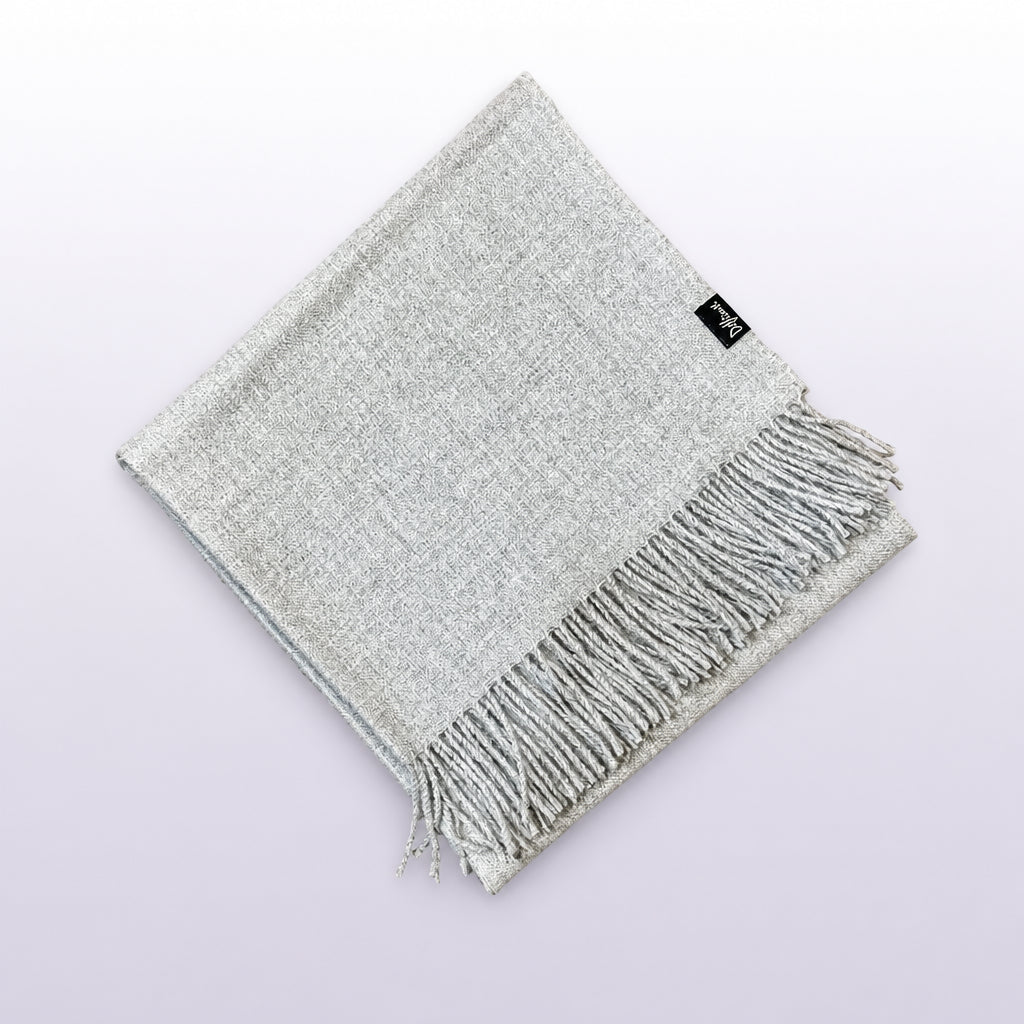 A folded light gray woven scarf with fringe on one end, placed on a rustic wooden surface. A small black brand tag is visible near the top edge.