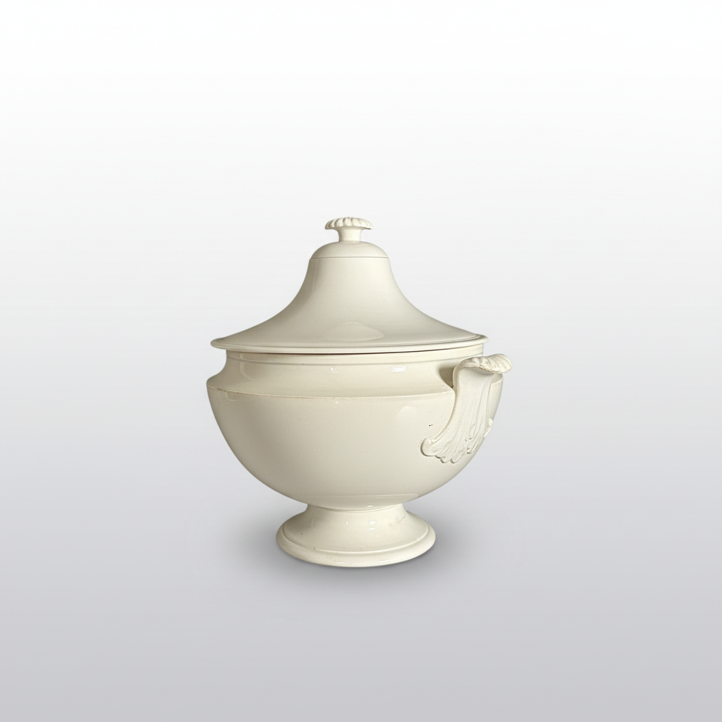 A white ceramic lidded serving bowl with decorative side handles and a pedestal base, photographed against a dark gray backdrop.