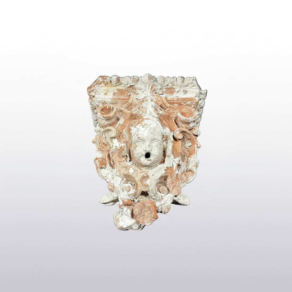 An ornate carved stone or plaster decorative wall piece featuring a central face surrounded by elaborate scrollwork and floral motifs, shown against a plain white background.