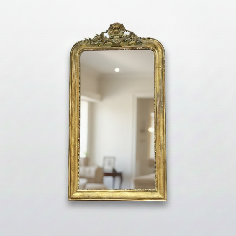 An ornate rectangular wall mirror with a distressed gold frame, mounted on a cream-colored wall in what appears to be an antique shop or gallery. The mirror is heavily cracked and reflects the store interior, including ceiling lights.