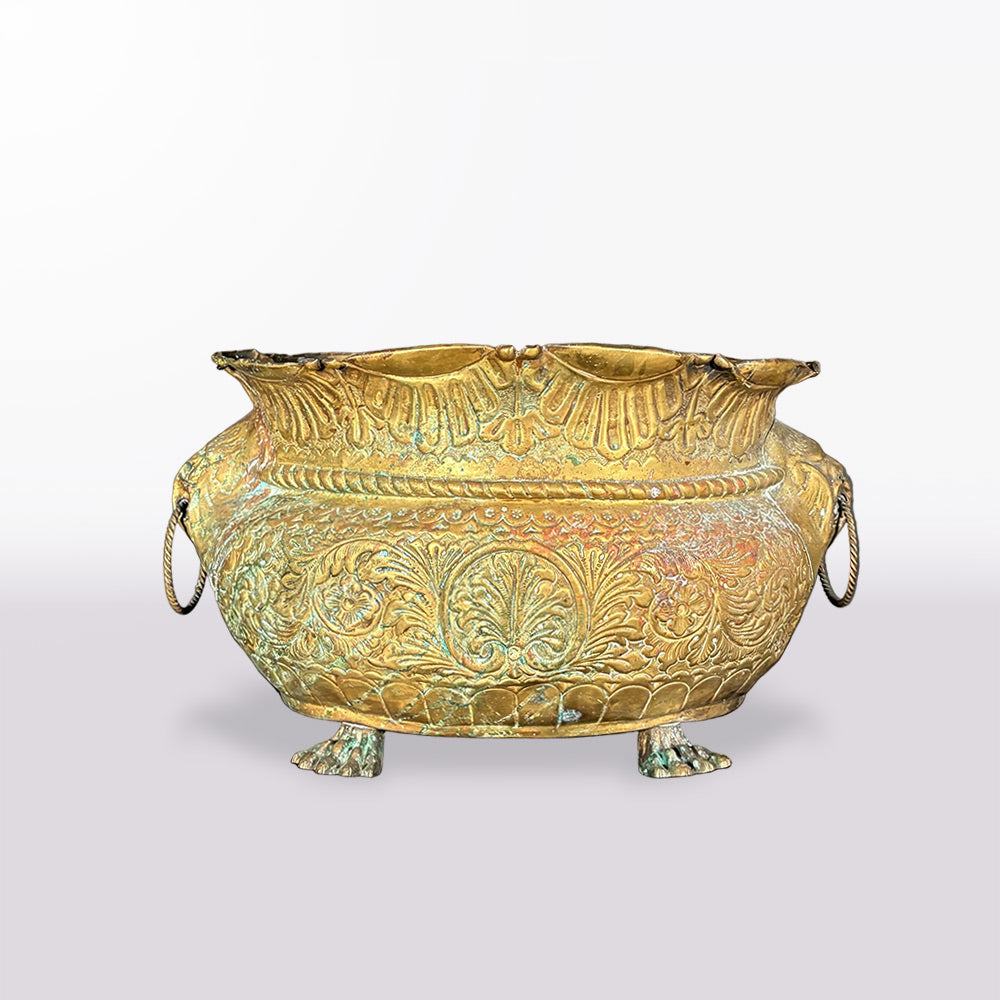 An ornate antique-style bronze or gold-toned metal cauldron with embossed floral relief patterns, rope handles on the sides, and three short legs, shown on a plain light gray background.