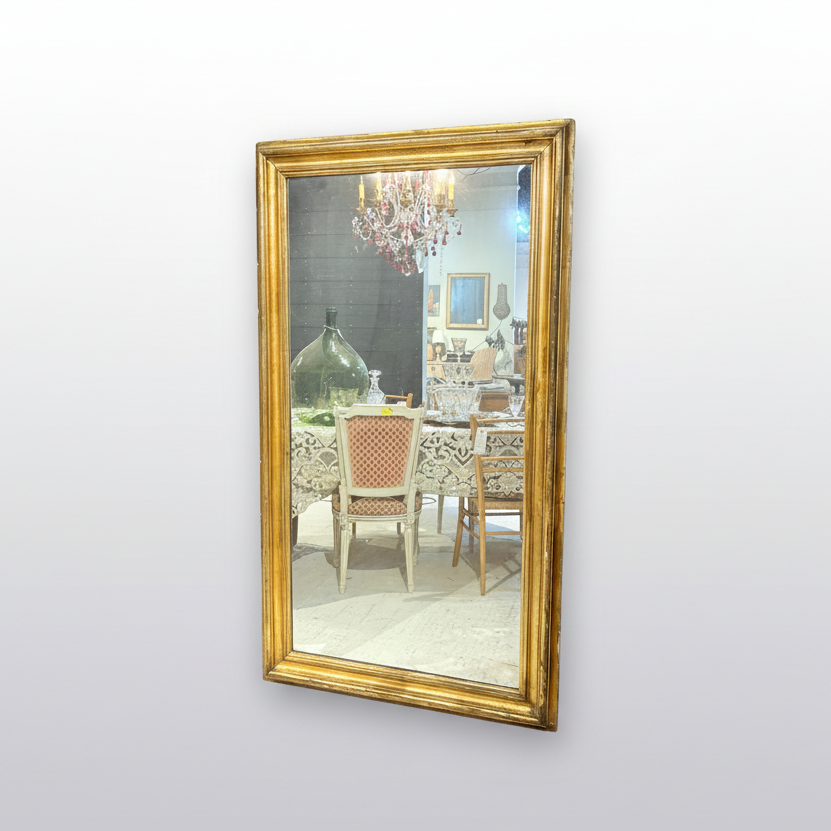 A large rectangular wall mirror with an ornate gold frame, leaning against a wall in a vintage furniture or decor store. The mirror reflects a shop interior with chairs and tables, and the surrounding area includes framed wall art, a rustic wooden cabinet, and patterned upholstery.