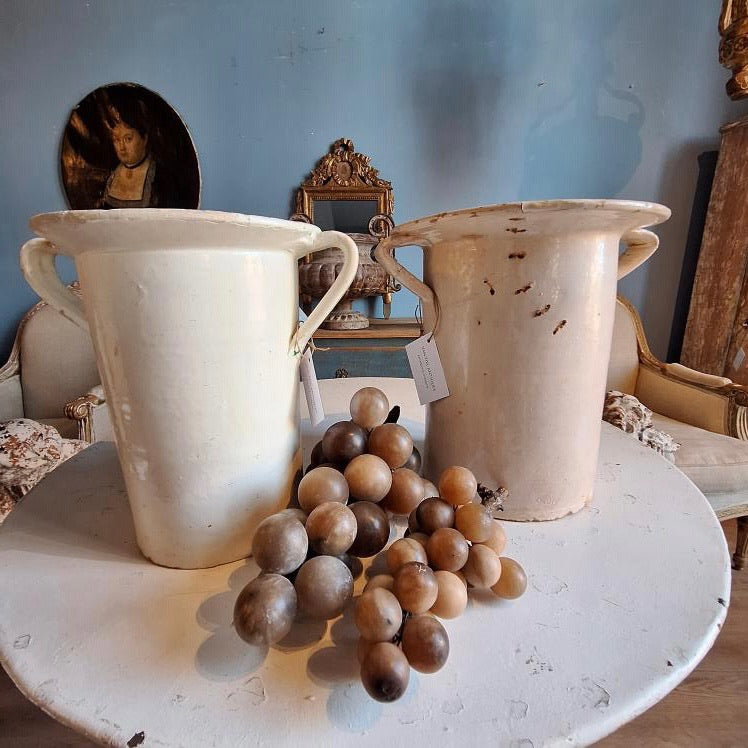 Two ceramic vases on a table with a cluster of grapes in front, against a decorative wall.