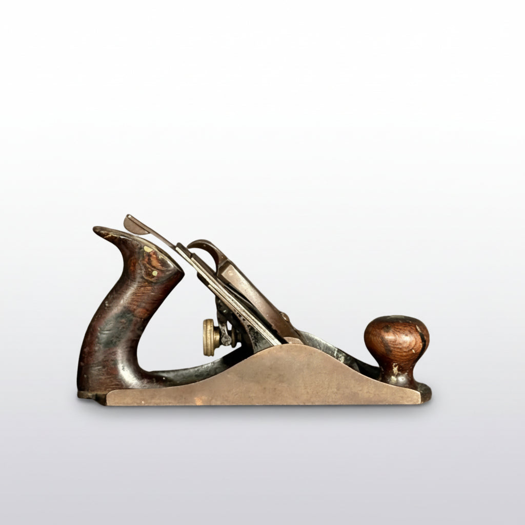 An antique wooden and metal hand plane with a dark brown wooden body, brass components, and a copper-toned knob, resting on a dark gray surface against a textured gray background.