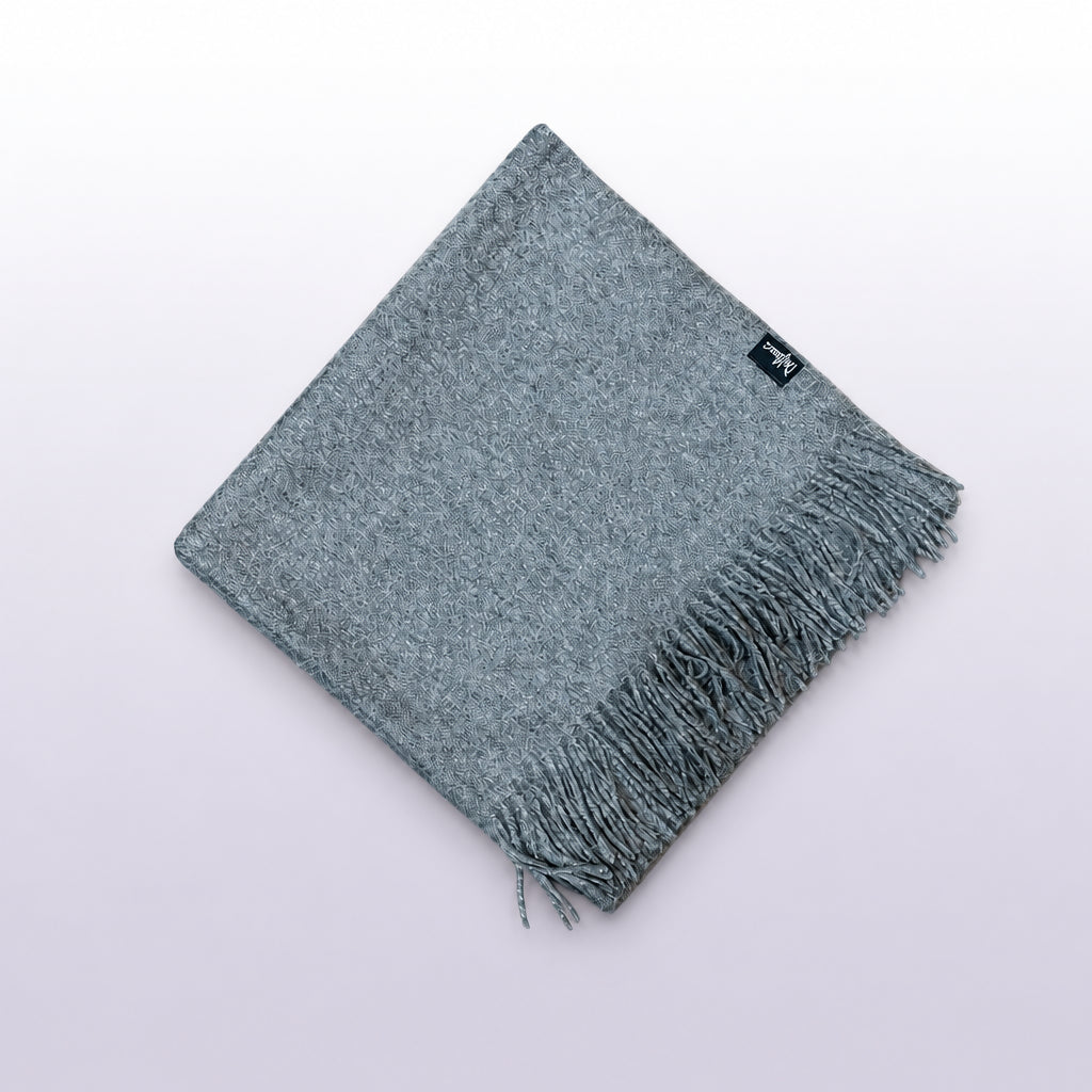 A folded gray knit scarf with fringe on one end, laid flat on a rustic wooden surface. A small black woven label is visible near the top edge.