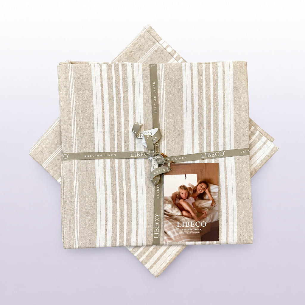 A folded Libeco Belgian Linen bedding set, featuring beige and white striped sheets or towels, wrapped in a matching ribbon with a small brand card, displayed on a rustic wooden background.